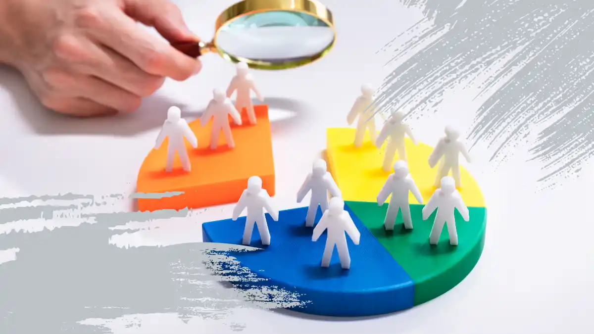 Hand holding a magnifying glass over a segmented pie chart with miniature human figures, representing customer segmentation and SEO-driven discovery marketing.